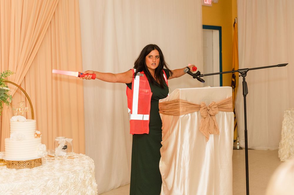Woman in black dress with red sash holds pink baton at wedding reception podium near white cake.