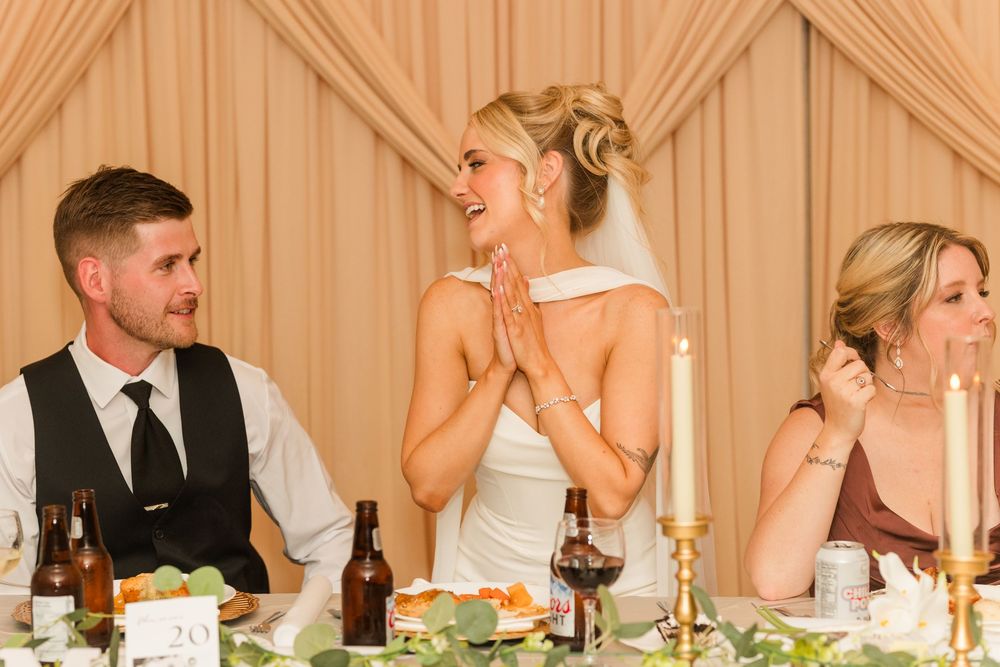 Bride laughing joyfully at wedding reception table with groom beside her.