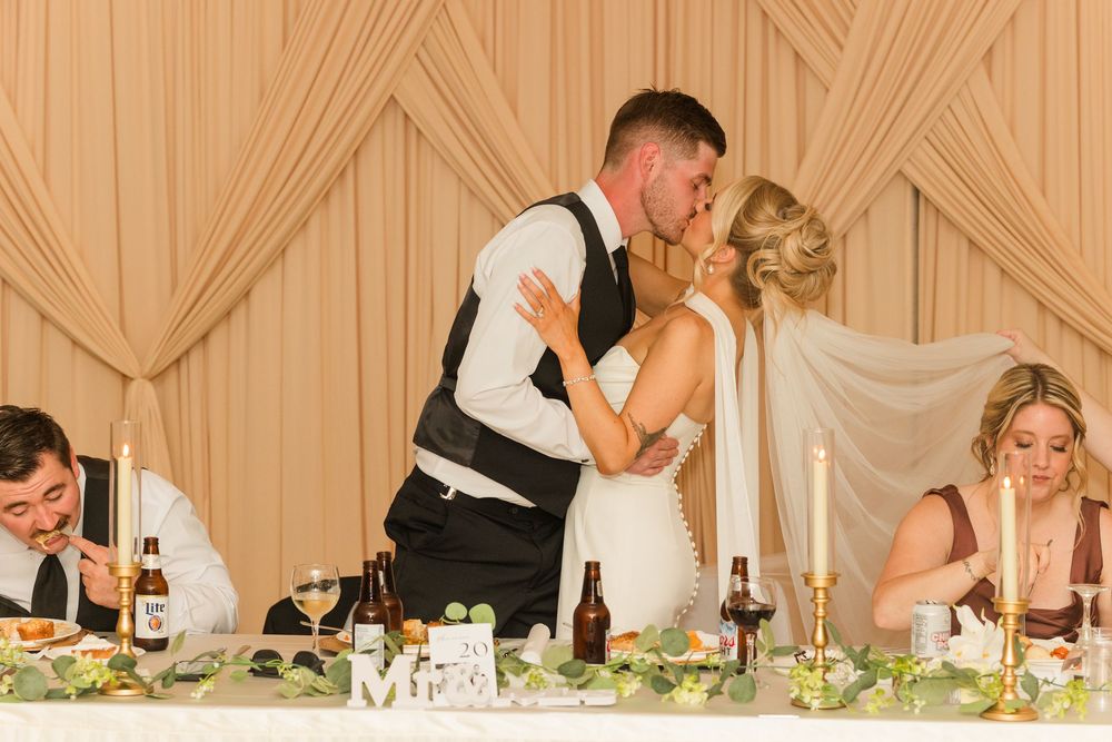 Bride and groom share a romantic kiss at wedding reception head table with candles and floral decor.