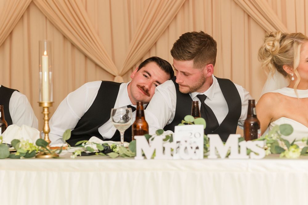 Two groomsmen laughing together at a wedding reception table decorated with Mr. and Mrs. signs and greenery.