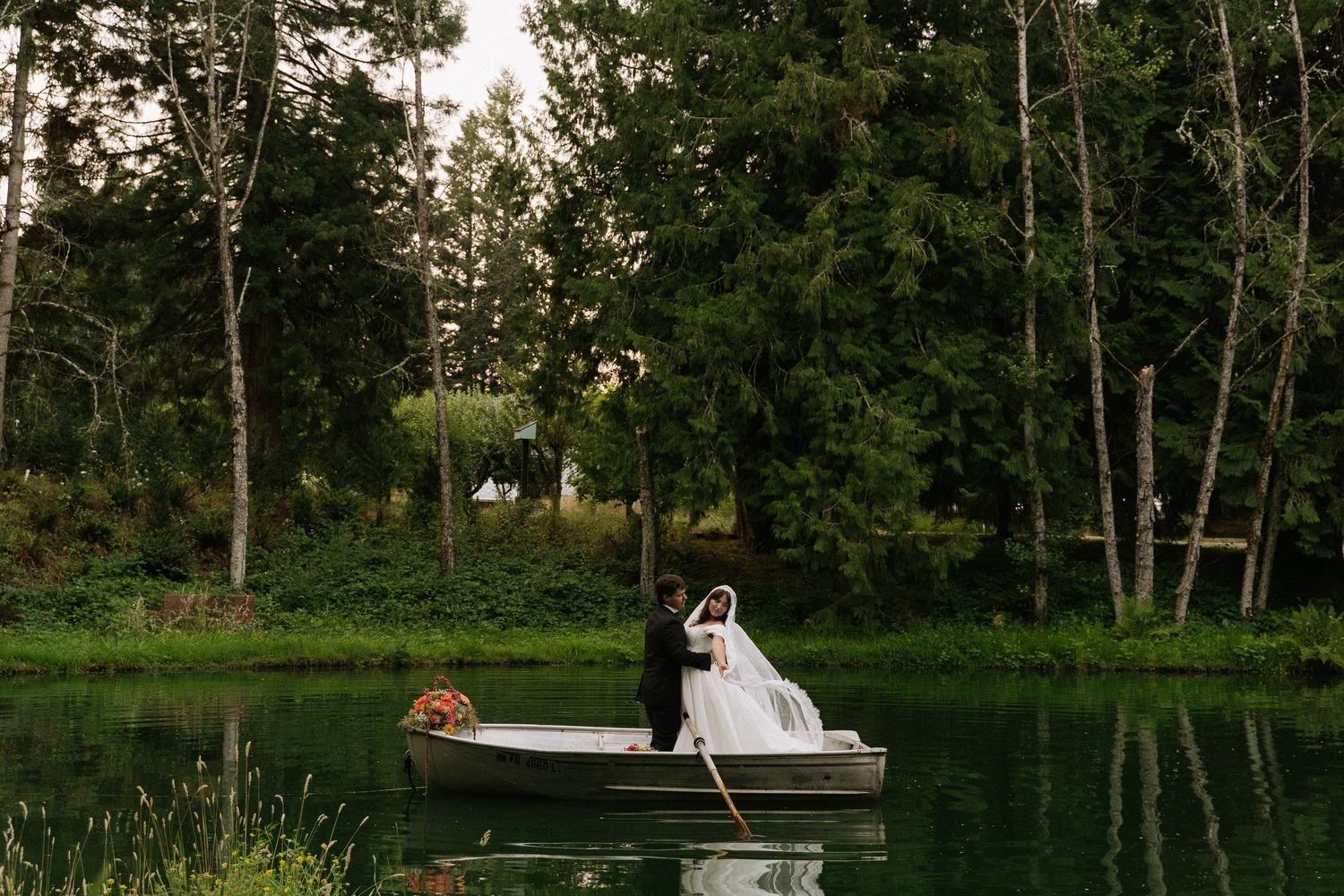 Bride and groom sharing a romantic moment on a rowboat surrounded by lush green trees on a serene lake.
