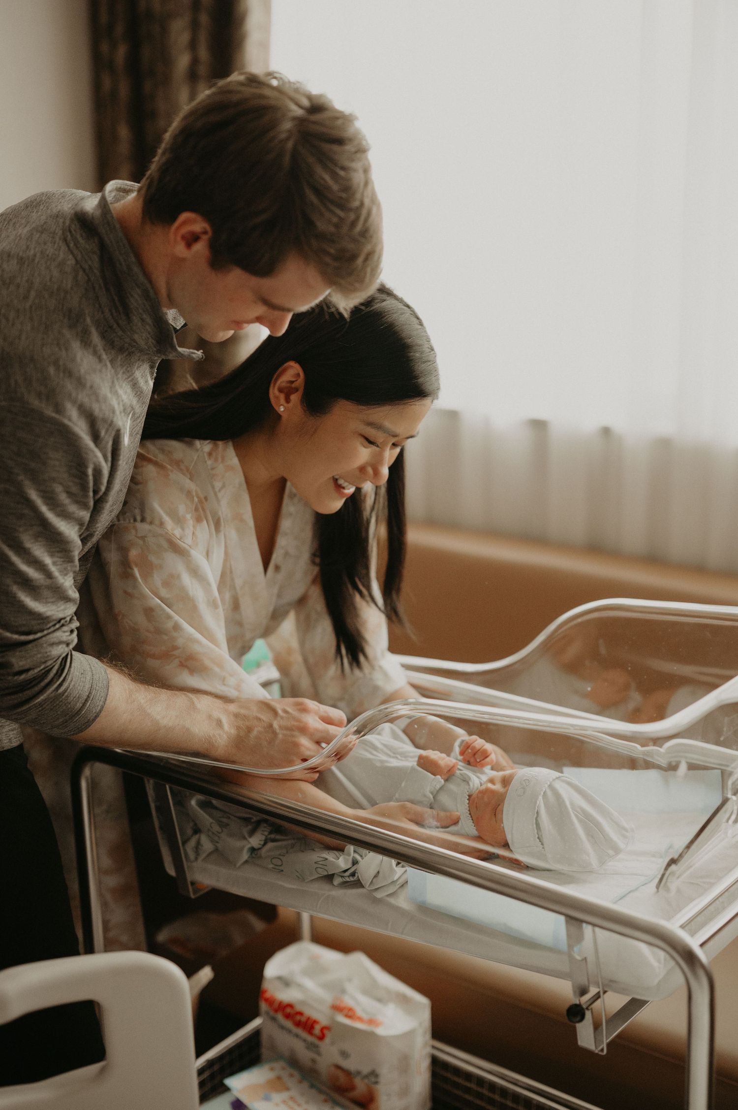 New parents smile lovingly at their newborn baby sleeping in a hospital bassinet in a bright, cozy maternity room.