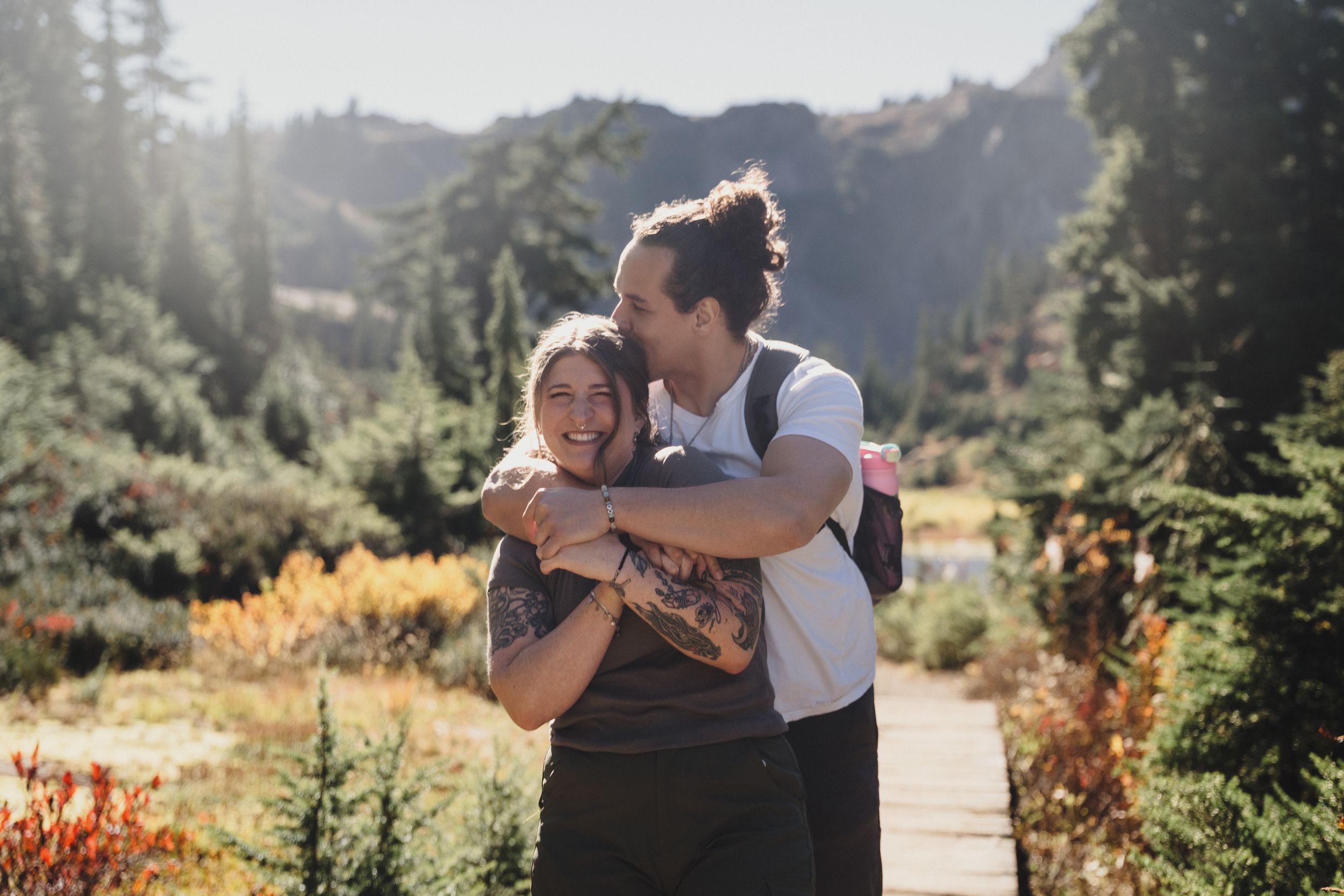 Smiling couple hugging on a scenic mountain trail surrounded by evergreen trees and colorful autumn foliage on a sunny day.