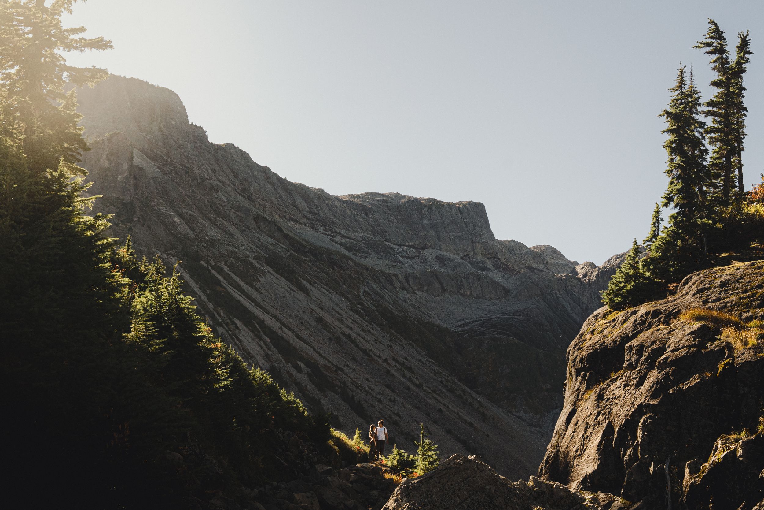 Golden sunlight illuminates a steep rocky mountain ridge with evergreen trees and a boulder-strewn foreground in a rugged alpine landscape.