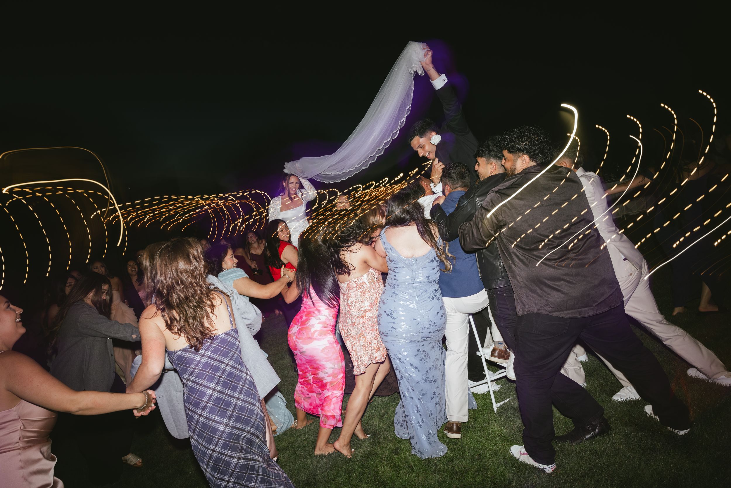 Guests dancing outdoors at night under string lights at a lively wedding reception celebration.