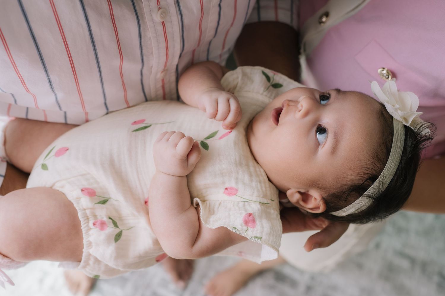 Newborn baby girl in floral romper and bow headband lying in mothers arms looking up curiously.