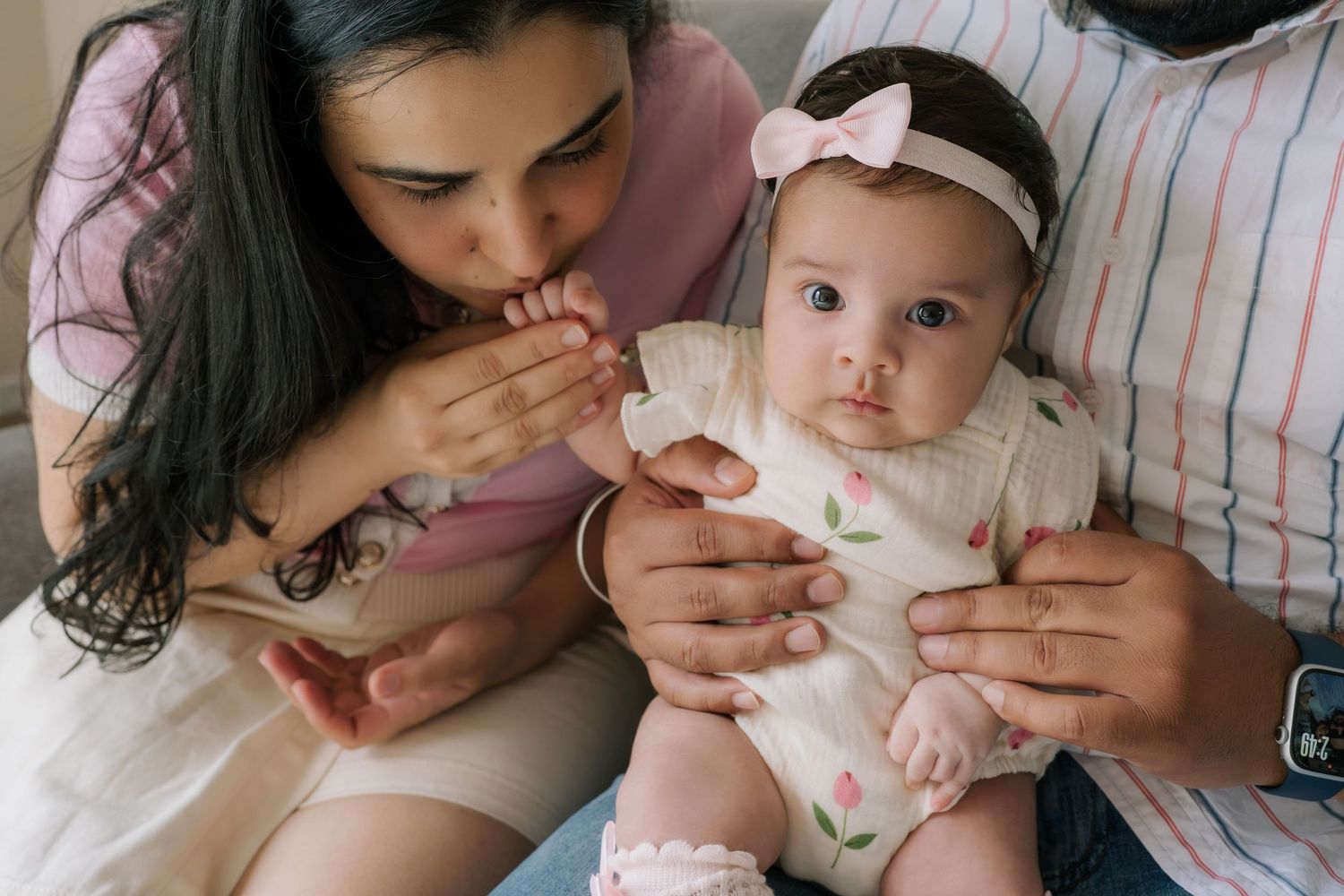 A mother kisses her baby girl's hand while the father holds the infant dressed in a floral outfit with a pink headband.