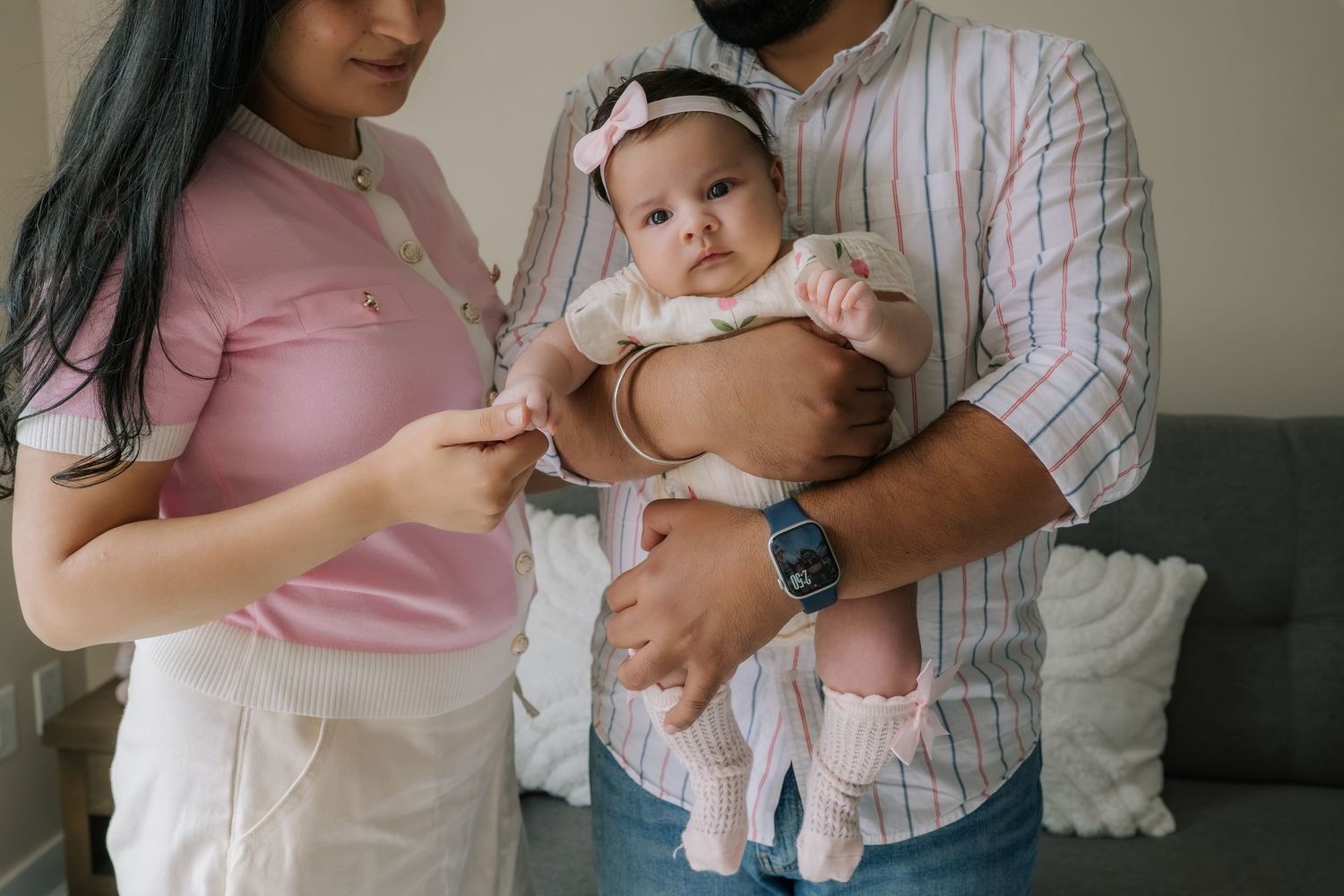 Parents holding their newborn baby girl in pink outfit and headband in a cozy home setting.