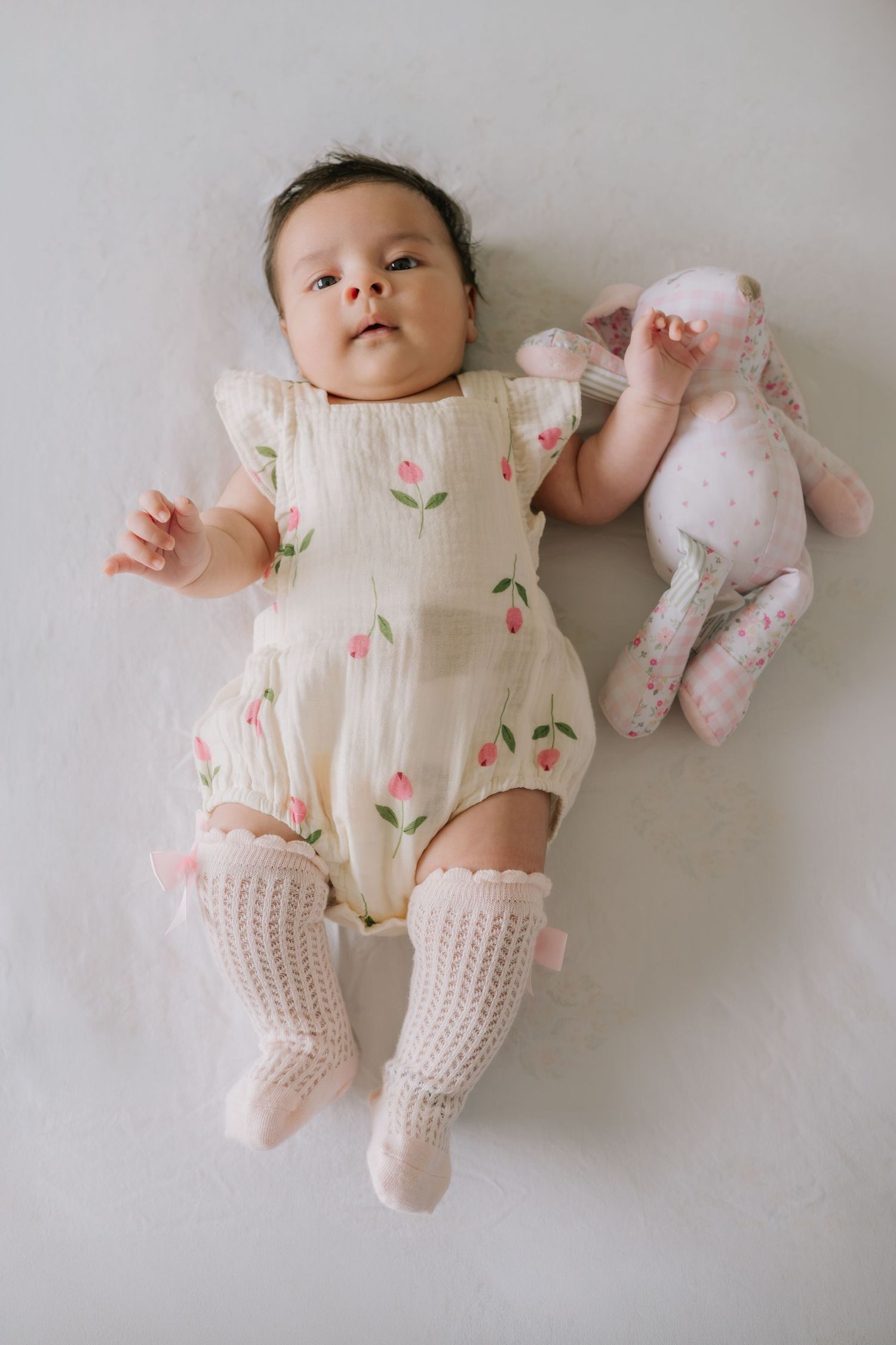 Baby girl in floral romper and pink knit socks lying next to a stuffed bunny toy on white background.