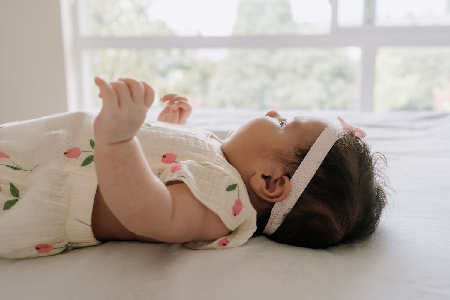 A newborn baby in a floral onesie and white headband lies on a bed, gazing upward with tiny hand raised.