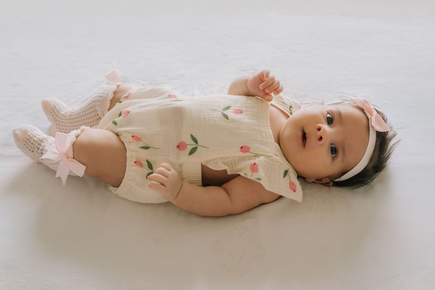 Baby girl in floral romper with pink headband and ruffle socks lying on white background.