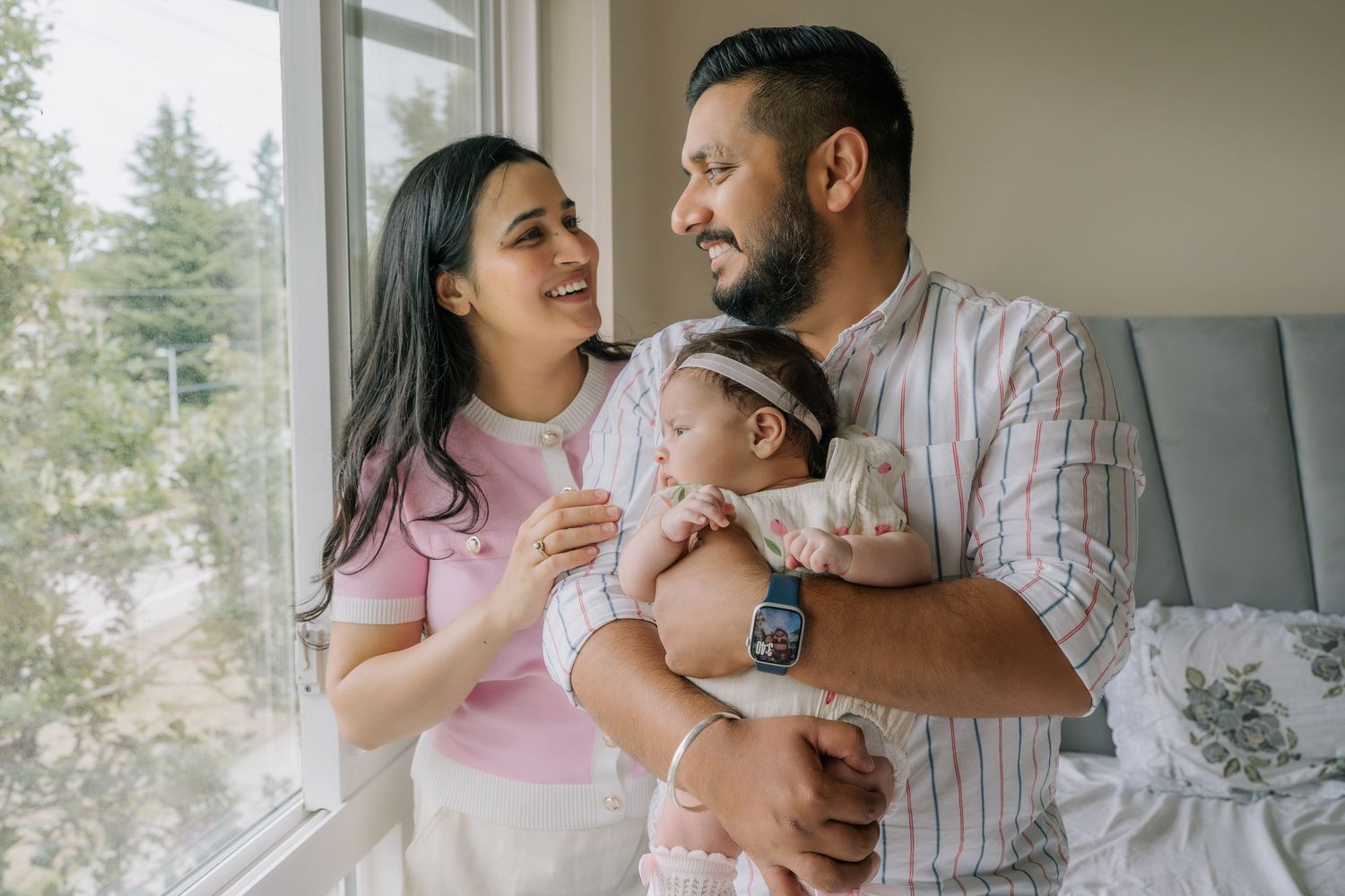 Happy young family with newborn baby standing by a sunlit window in a cozy bedroom at home.