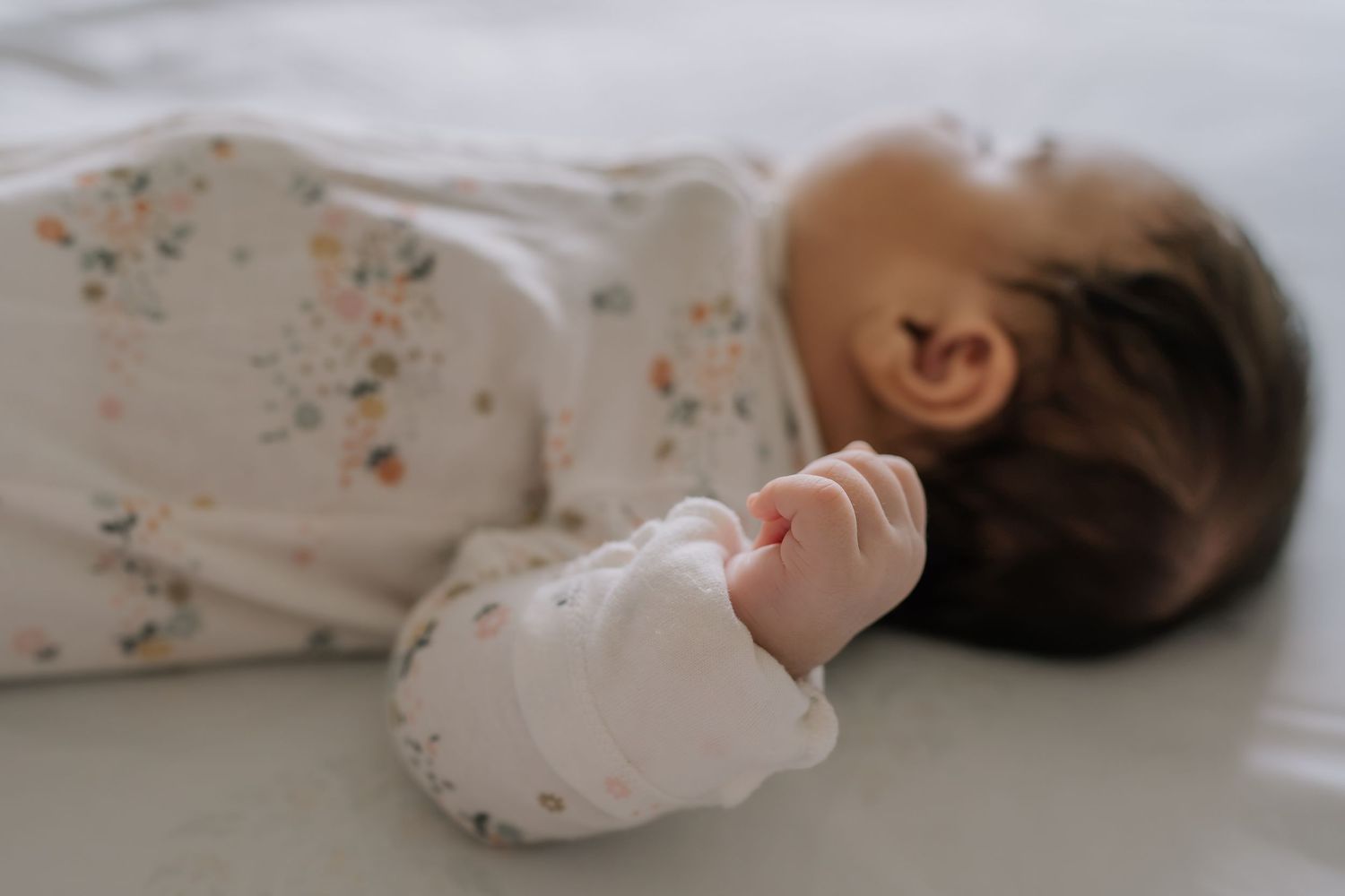 A sleeping newborn baby in a floral onesie lies peacefully on a white surface with tiny fists curled up.