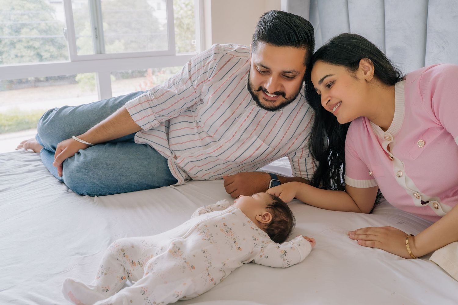 Happy parents lying on bed smiling at their newborn baby dressed in white, enjoying a tender family moment.
