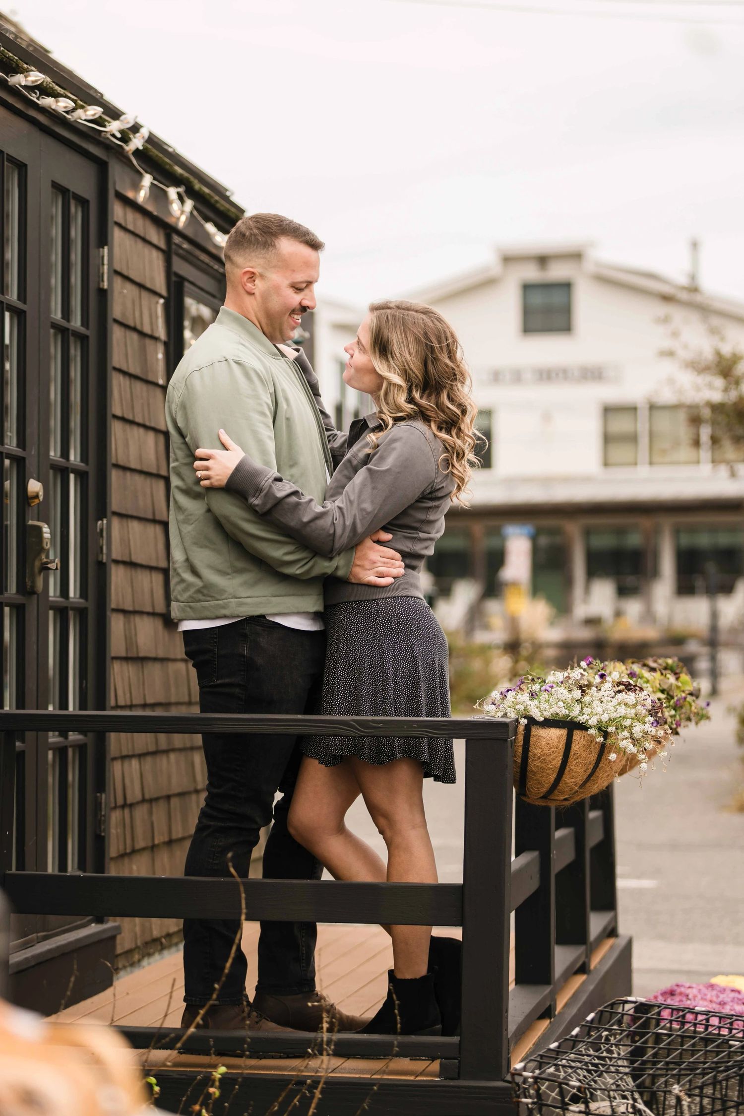 Couple embracing on a wooden deck outside a residential building, looking at each other intimately. Engagement at Rockport Harbor, Rockport
