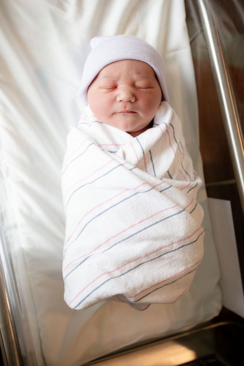 Peaceful newborn baby in a lavender knit cap, swaddled snugly in a pink and blue striped hospital blanket while sleeping in a bassinet.