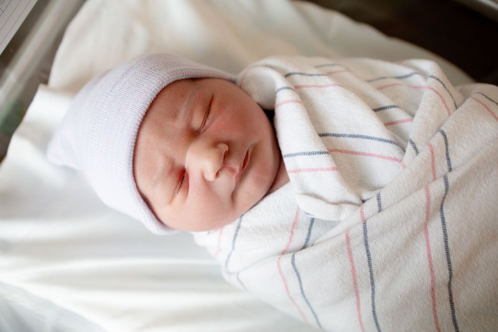 Sleeping newborn baby in a white knit hat, swaddled in a pink and blue striped hospital blanket.