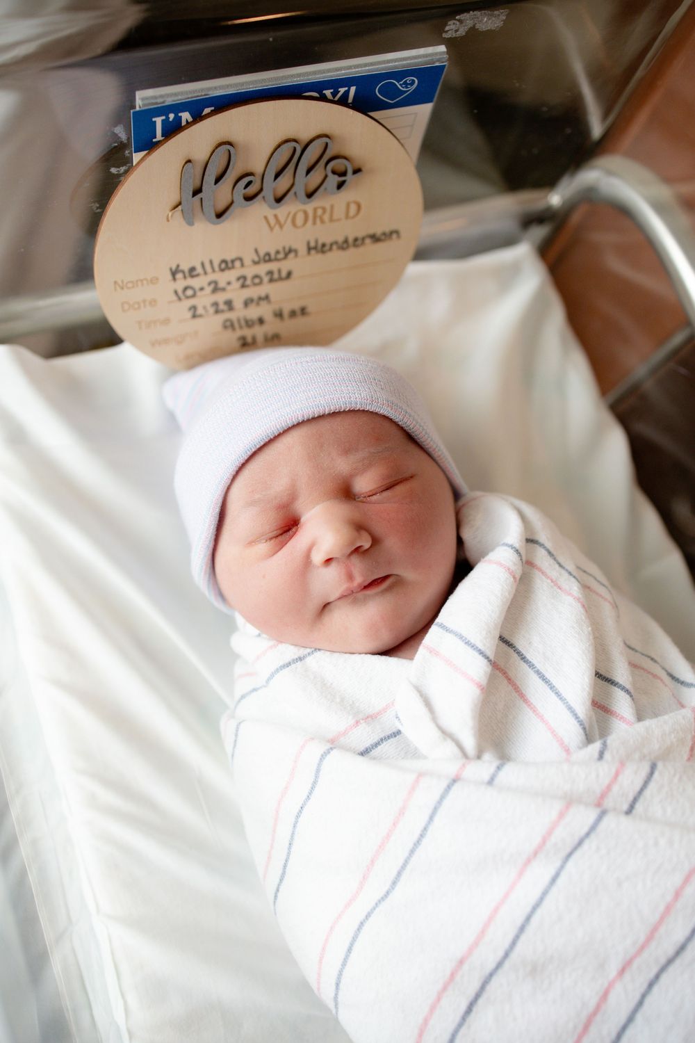 Sleeping newborn in a white knit hat swaddled in a striped hospital blanket beneath a wooden Hello World birth announcement sign.