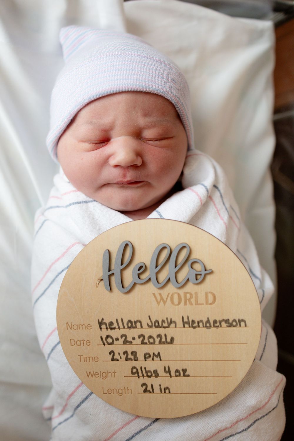 Sleeping newborn in a white knit cap and striped blanket beside a Hello World birth announcement sign listing name, date, weight and length.