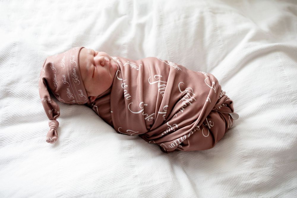 Newborn baby swaddled in a mauve patterned wrap and matching knotted hat, sleeping peacefully on soft white bedding.