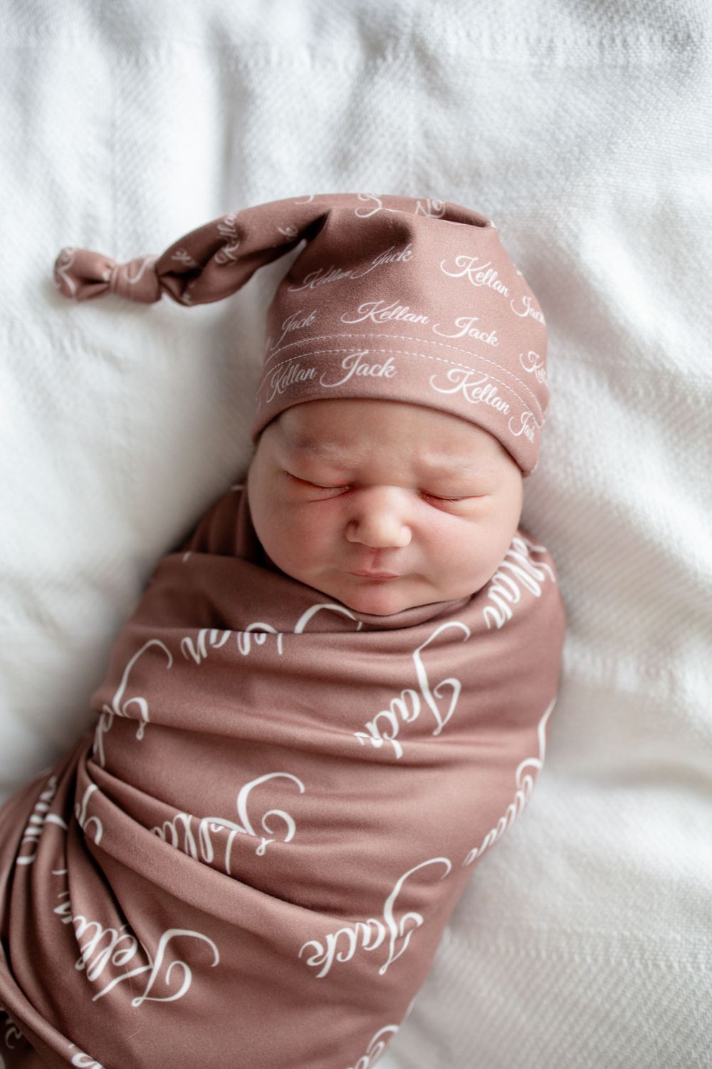 Newborn baby asleep in a mauve swaddle and knotted hat personalized with the name Kellan Jack in white script lettering.