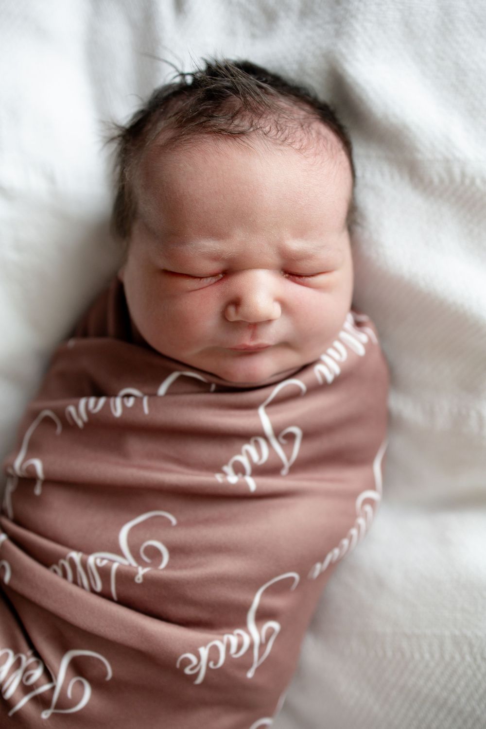 Sleeping newborn baby swaddled in a dusty rose blanket with white script lettering, resting peacefully on a soft white background.