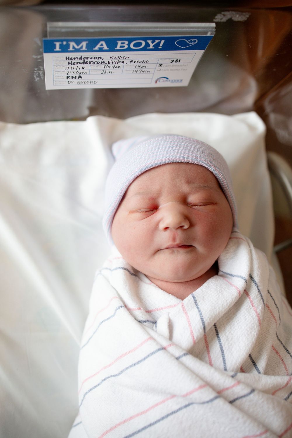 Sleeping newborn baby boy in a white knit hat, swaddled in a striped hospital blanket, resting in a bassinet beneath an I'm a Boy card.