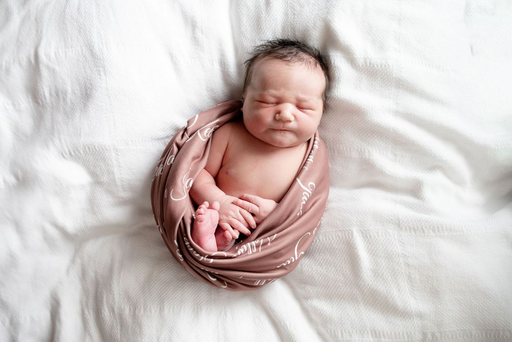 Sleeping newborn baby wrapped in a dusty rose swaddle with a leaf pattern, resting on a soft white blanket.