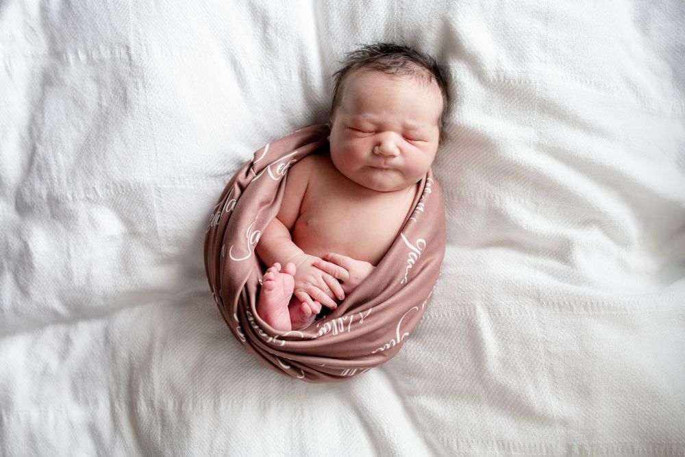 Sleeping newborn baby wrapped in a mauve patterned swaddle, lying peacefully on soft white bedding in a newborn photography session.