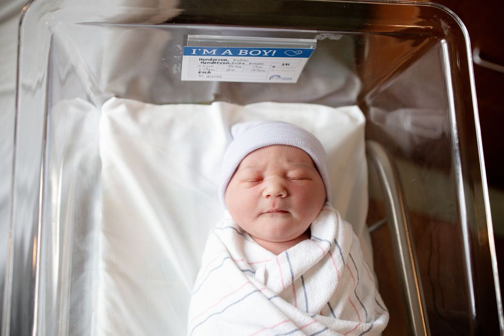 Sleeping newborn baby in a white knit hat, swaddled in a striped blanket inside a hospital bassinet with an "I'm a boy" card.