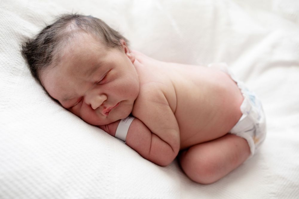 Sleeping newborn baby with dark hair and a hospital wristband rests on its side on soft white bedding wearing a diaper.