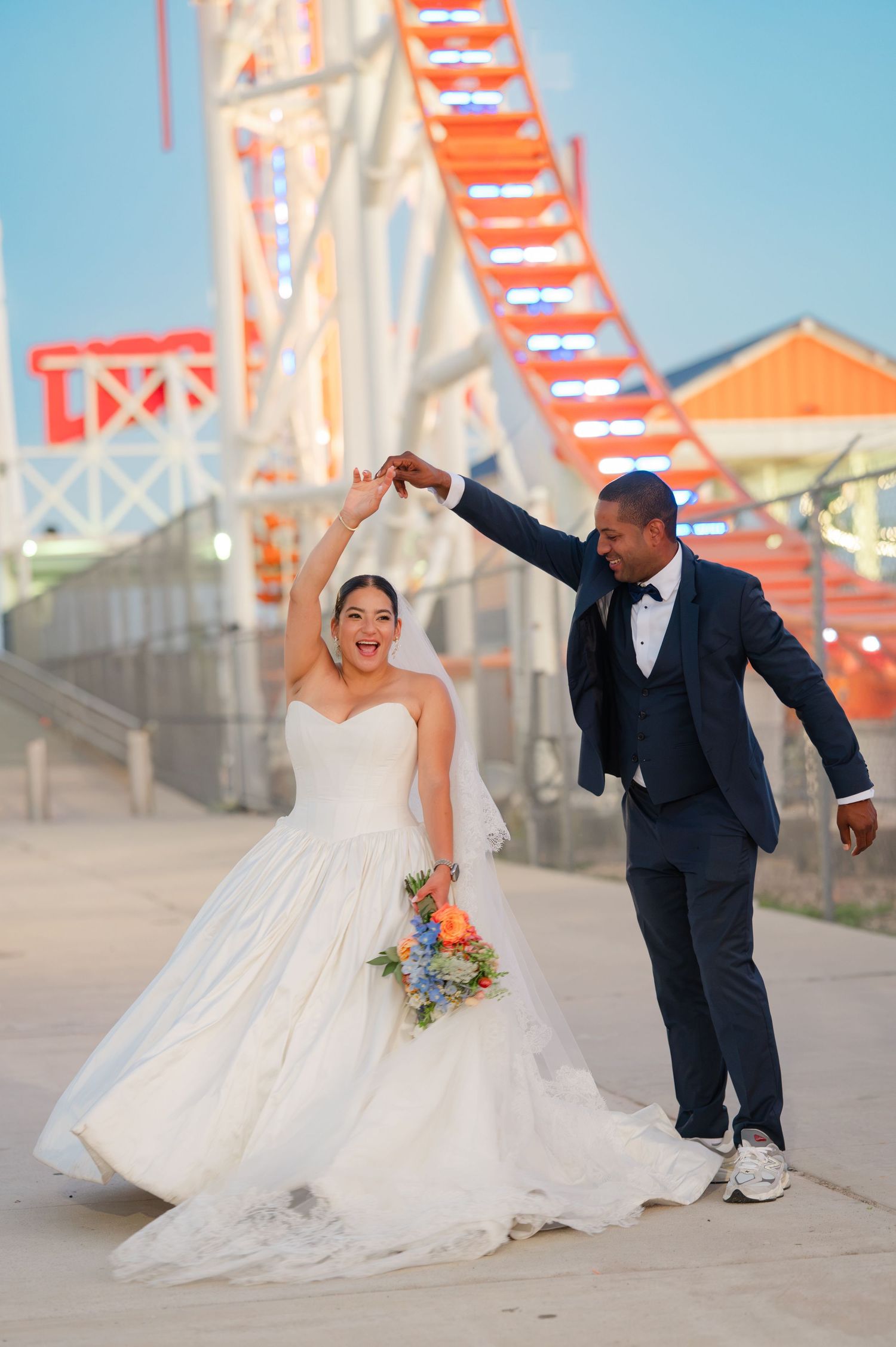 Joyful bride in a white strapless gown holding a  bouquet twirls with her groom in a navy suit in front of a roller coaster in Coney Island.