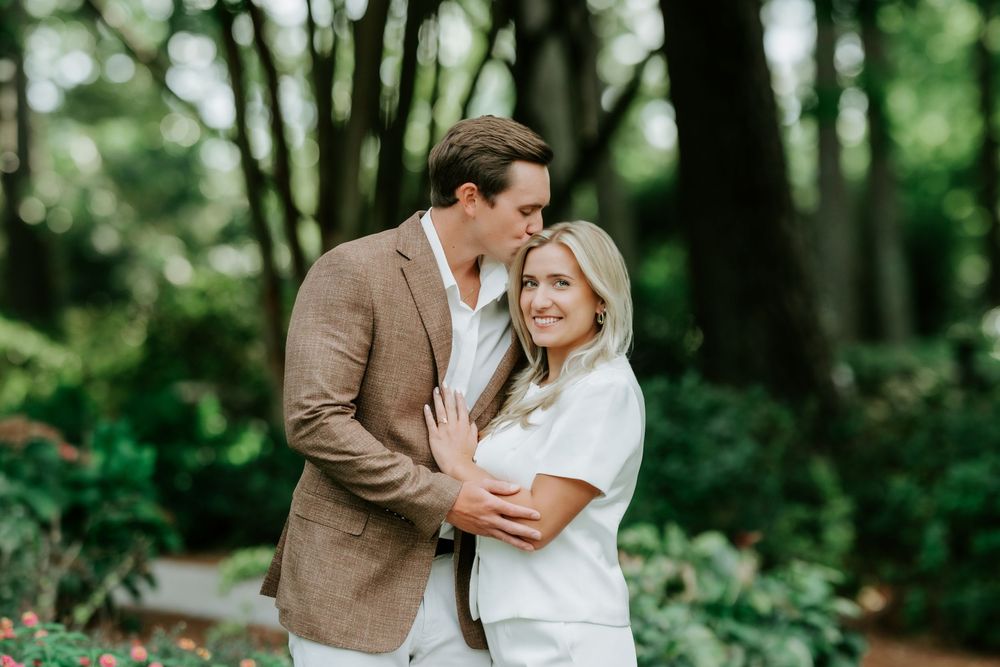 Couple embracing in lush green garden, man in tan blazer whispering to smiling blonde woman in white outfit.
