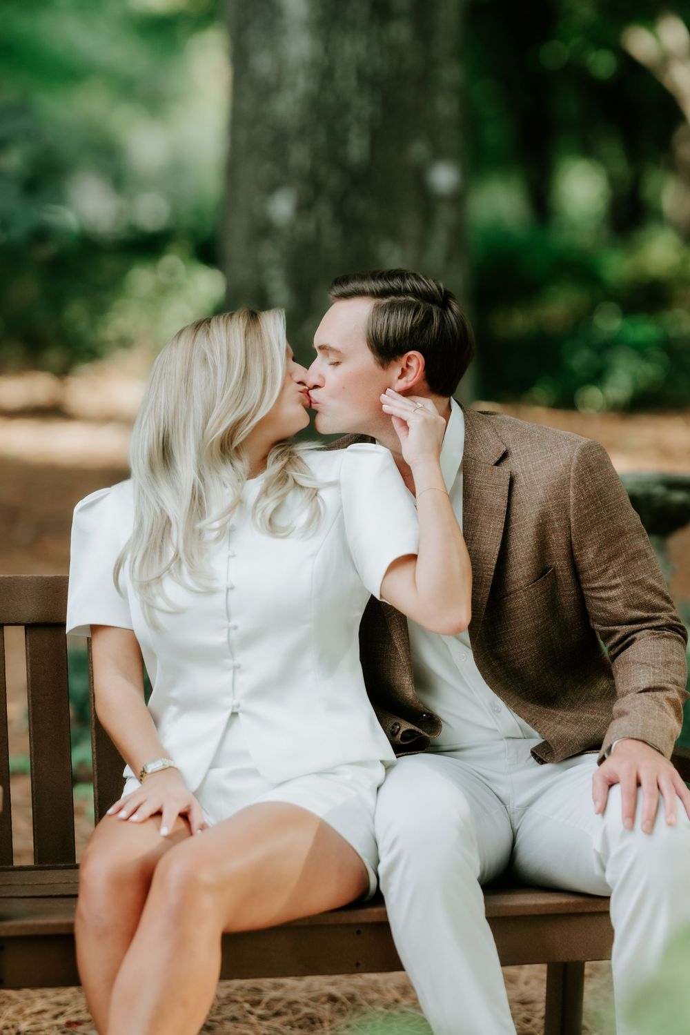 Couple sharing a romantic kiss on a wooden bench surrounded by lush green trees outdoors.