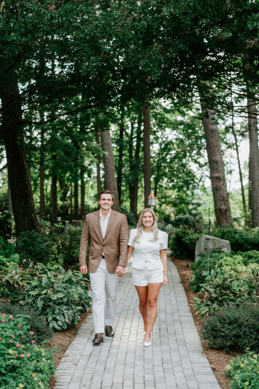 Couple holding hands walking down a brick garden path surrounded by lush green trees and flowering shrubs.