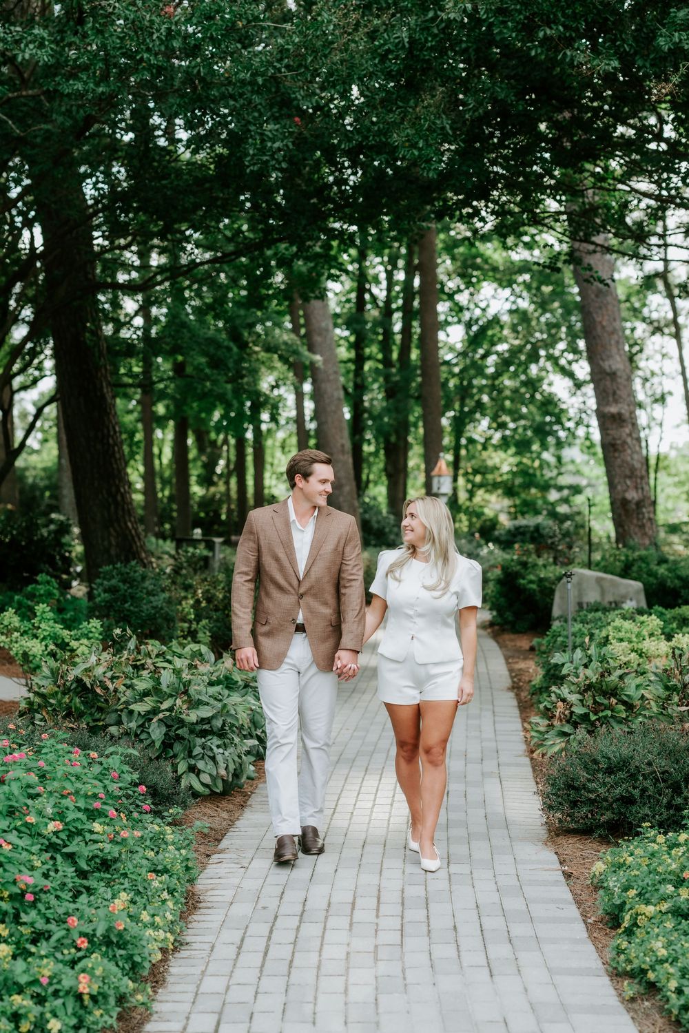 Couple holding hands walking down brick garden path, man in tan suit and woman in white romper smiling at each other.