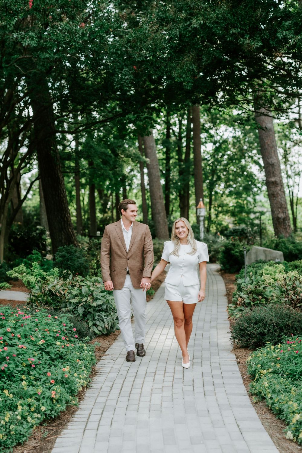 Couple holding hands walking along a brick garden path lined with trees and colorful flowers.