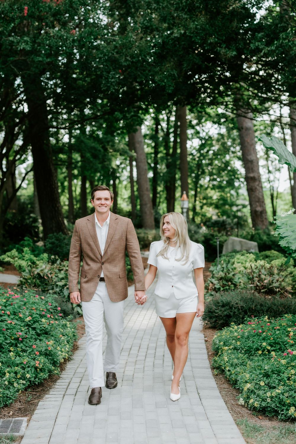 Couple holding hands walking on brick path in lush garden, man in tan blazer, woman in white romper.