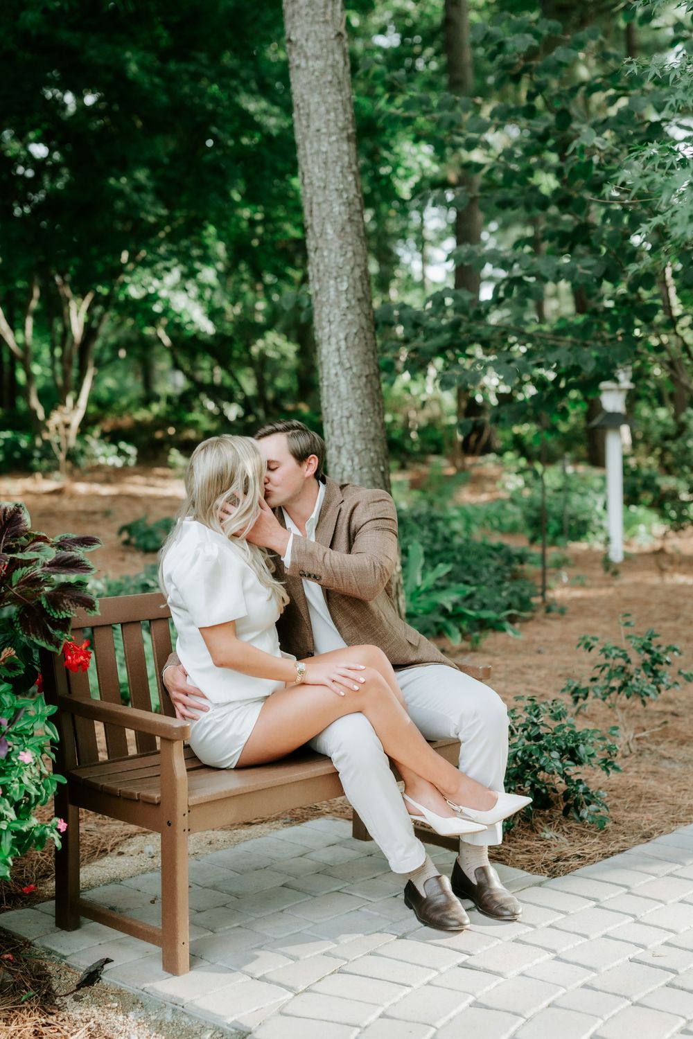 Couple kissing on a wooden garden bench surrounded by tall pine trees and green shrubs.