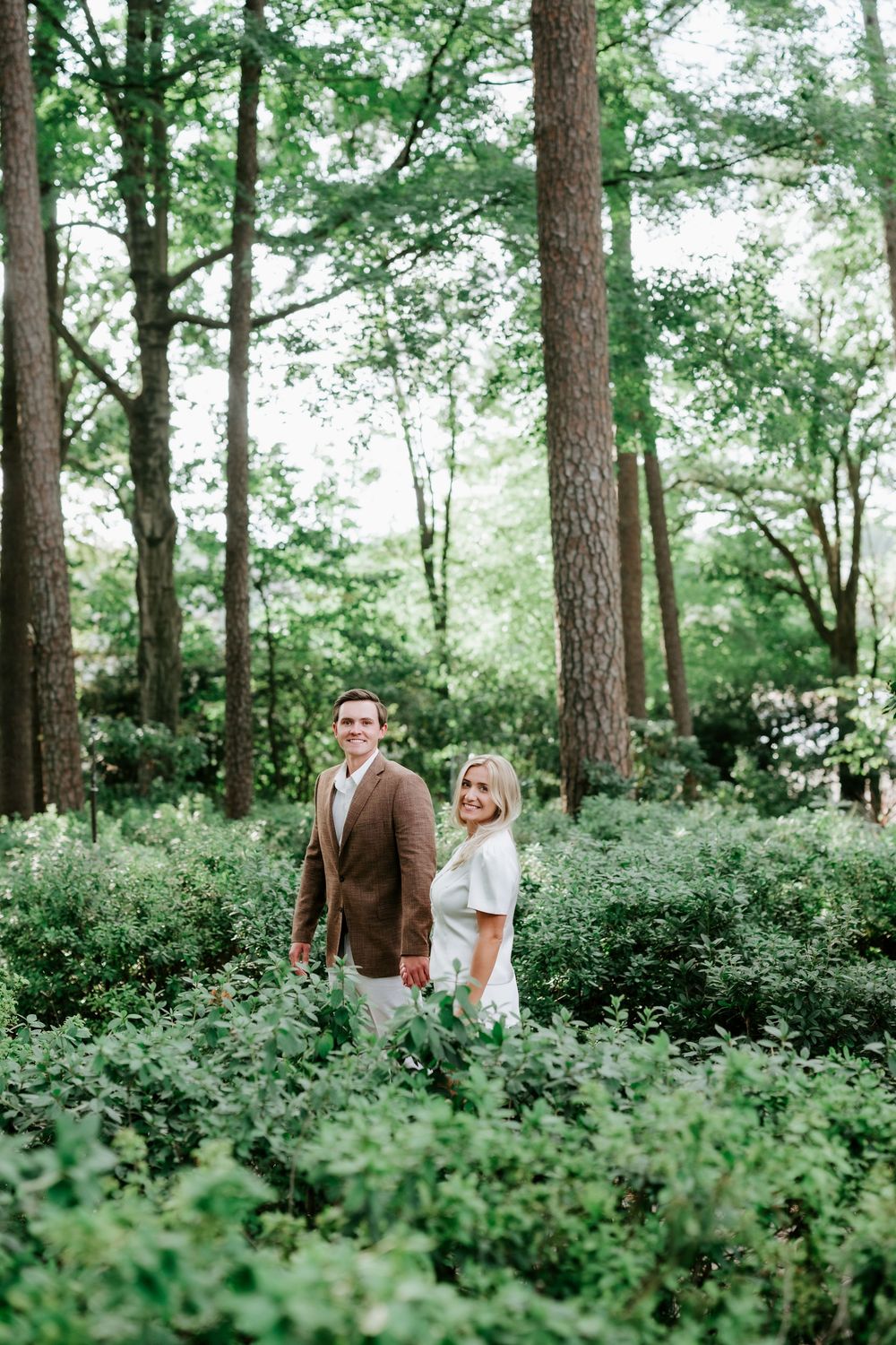 Couple smiling in lush green forest, man in brown blazer and woman in white dress holding hands.
