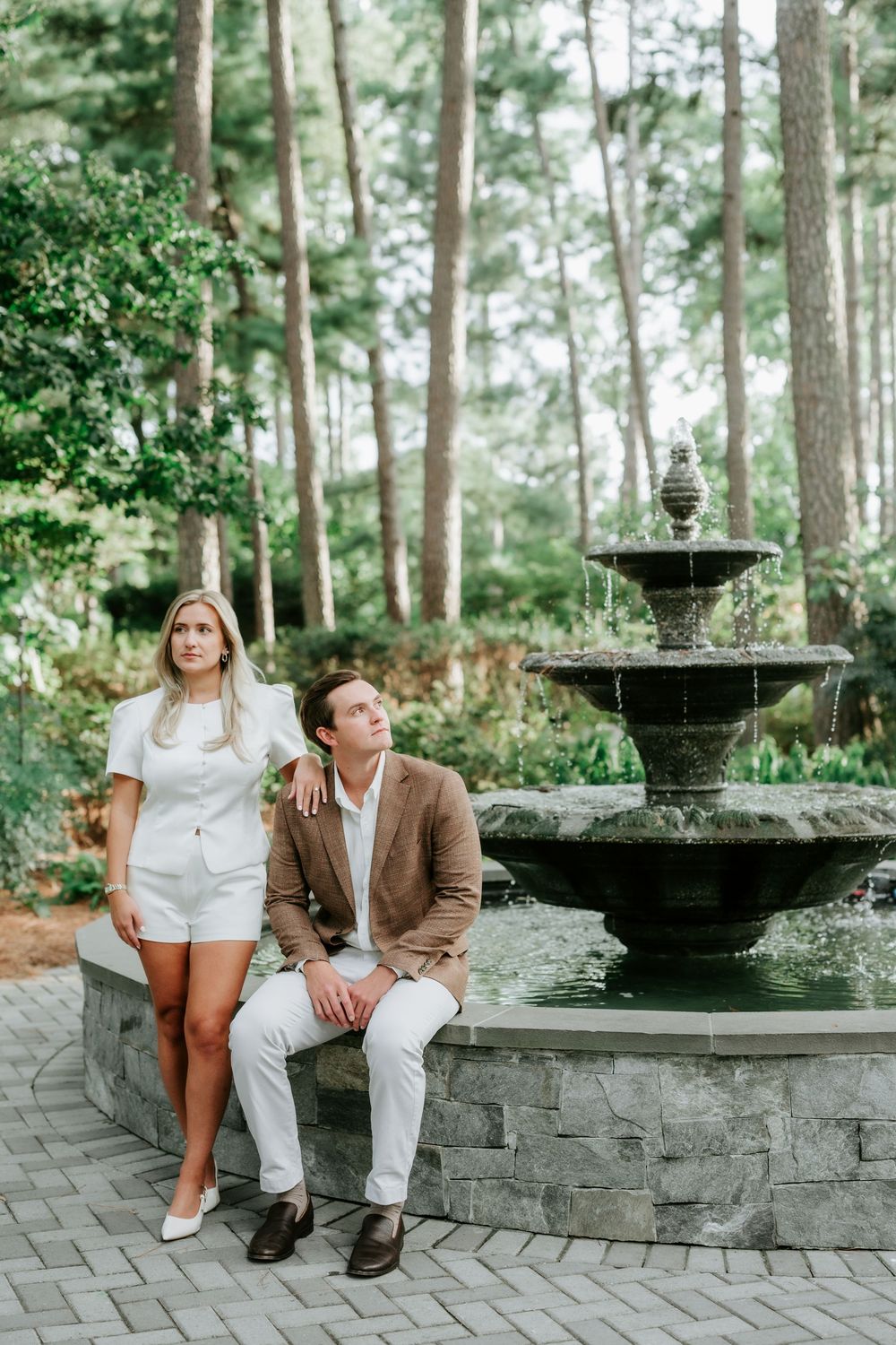 Couple posing by a stone tiered fountain surrounded by tall pine trees in an elegant garden setting.