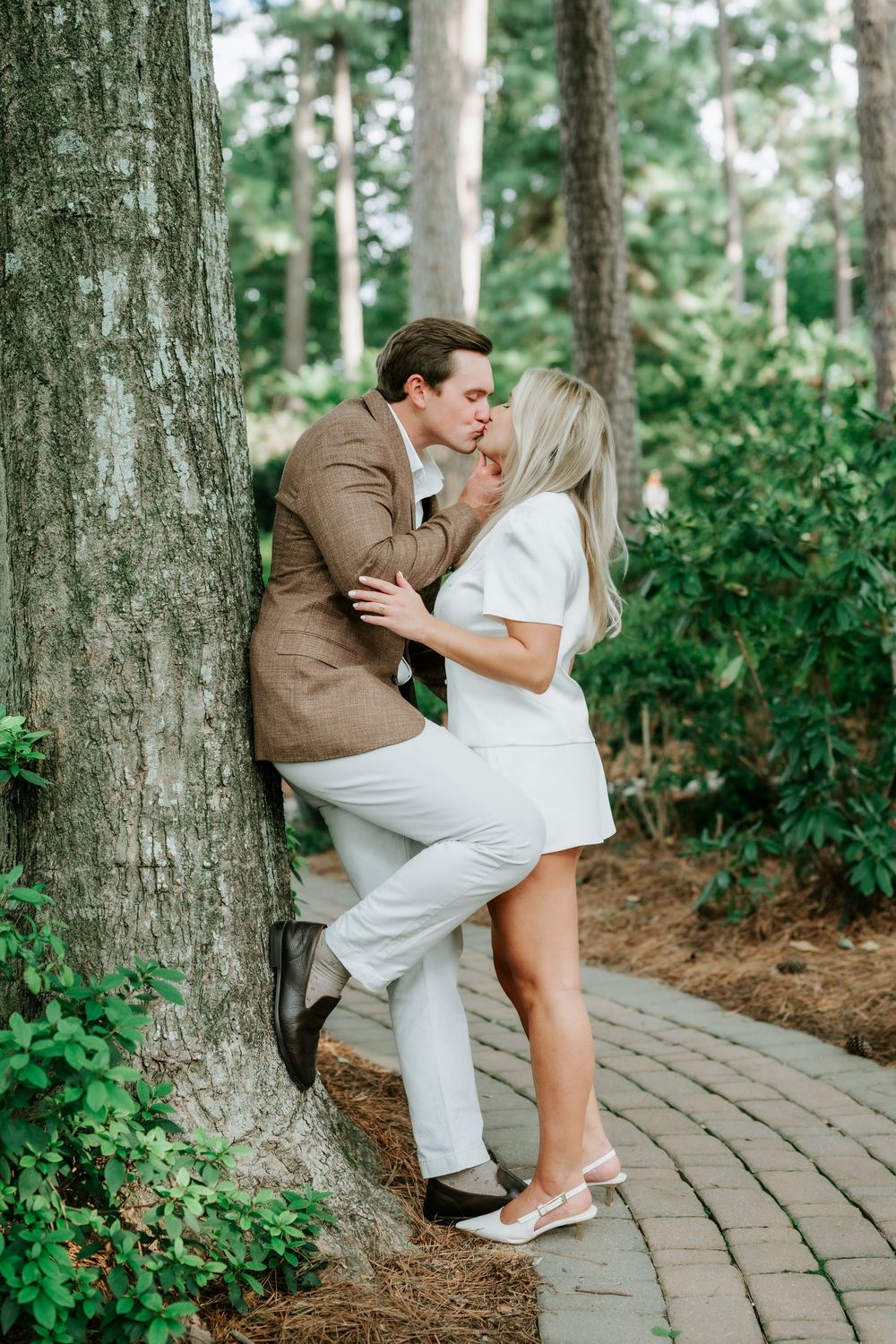 Couple sharing a passionate kiss while leaning against a large tree trunk in a garden setting.