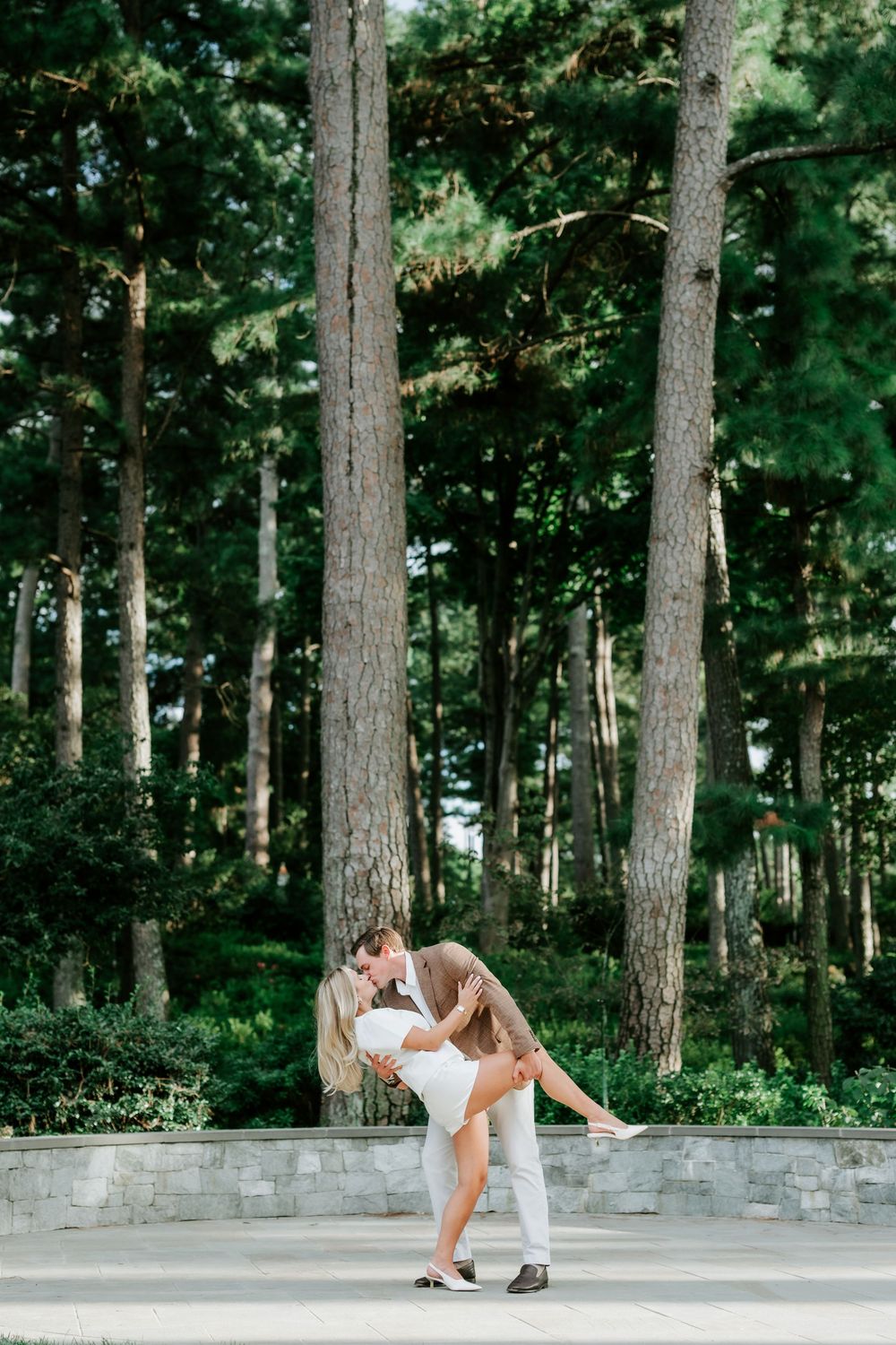 Couple sharing a romantic dip kiss in front of tall pine trees in a lush garden setting.