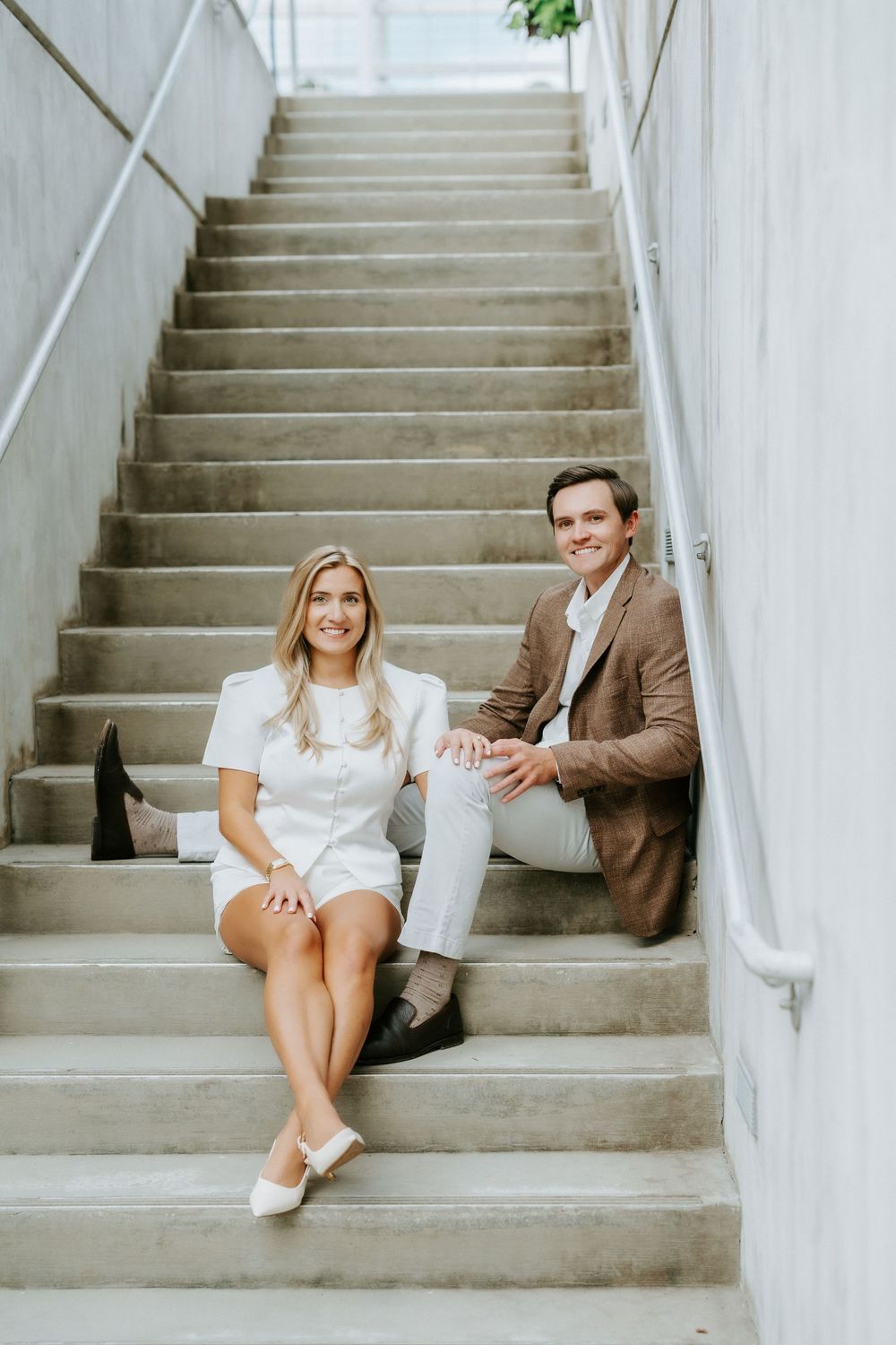 Couple sitting on concrete stairs, woman in white mini dress and man in brown blazer smiling.