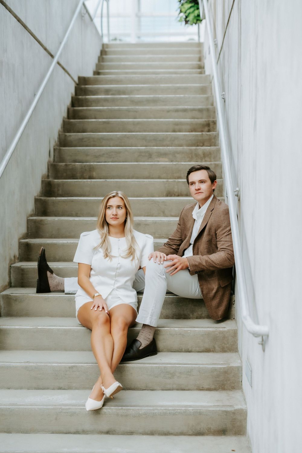 Couple posing seriously on concrete steps, woman in white dress and man in brown suit jacket.