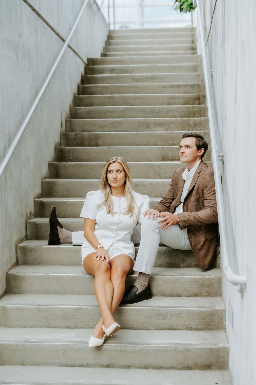 Couple sitting on modern concrete stairs, woman in white dress, man in brown blazer.
