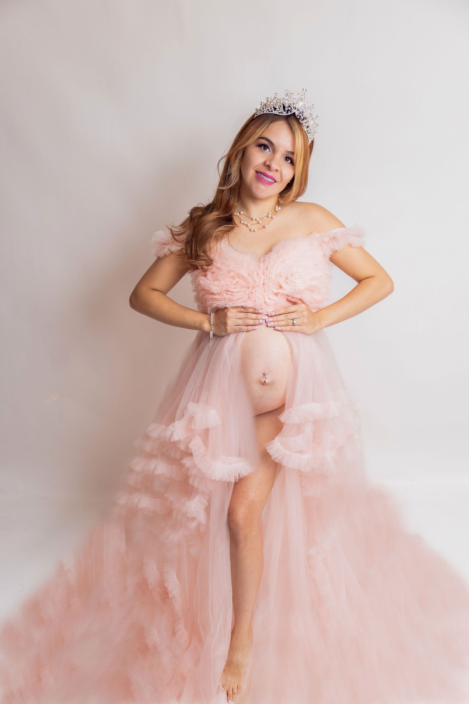 Pregnant woman posing in pink ruffled maternity gown with tiara and pearl necklace on white backdrop.