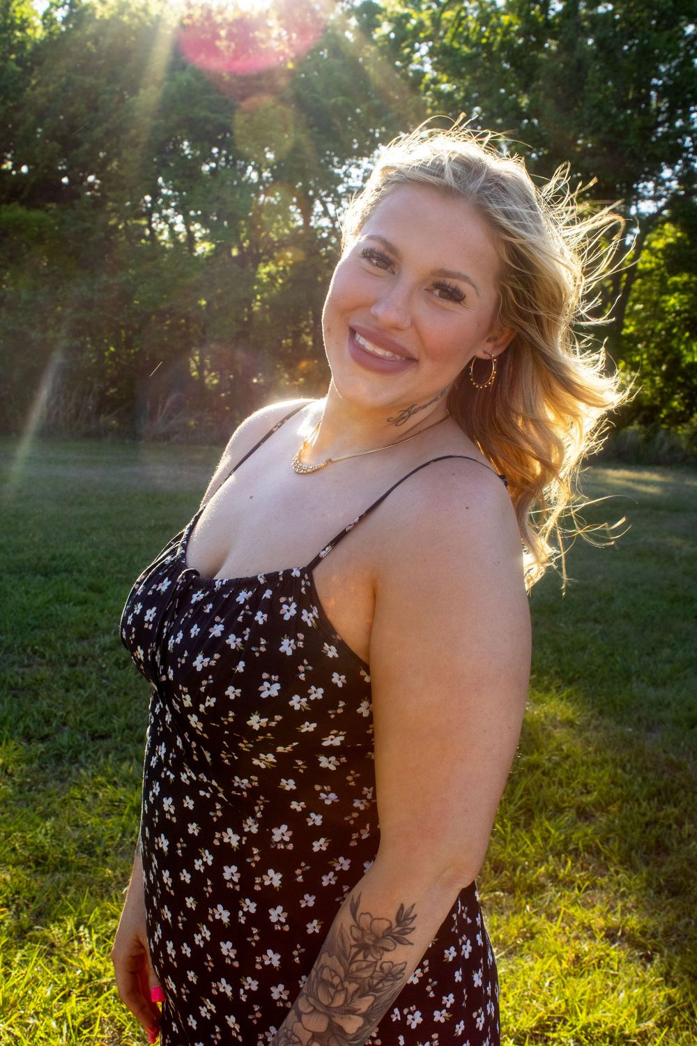 Smiling blonde woman in floral sundress with tattoo poses outdoors in golden sunlight with green trees behind her.