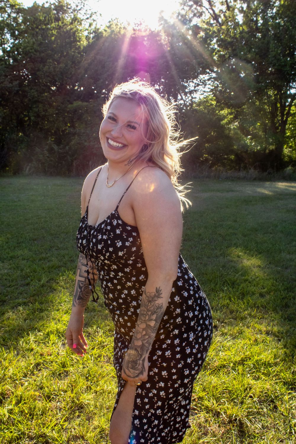 Smiling blonde woman with tattoos in floral sundress posing outdoors in sunlit park on sunny day.