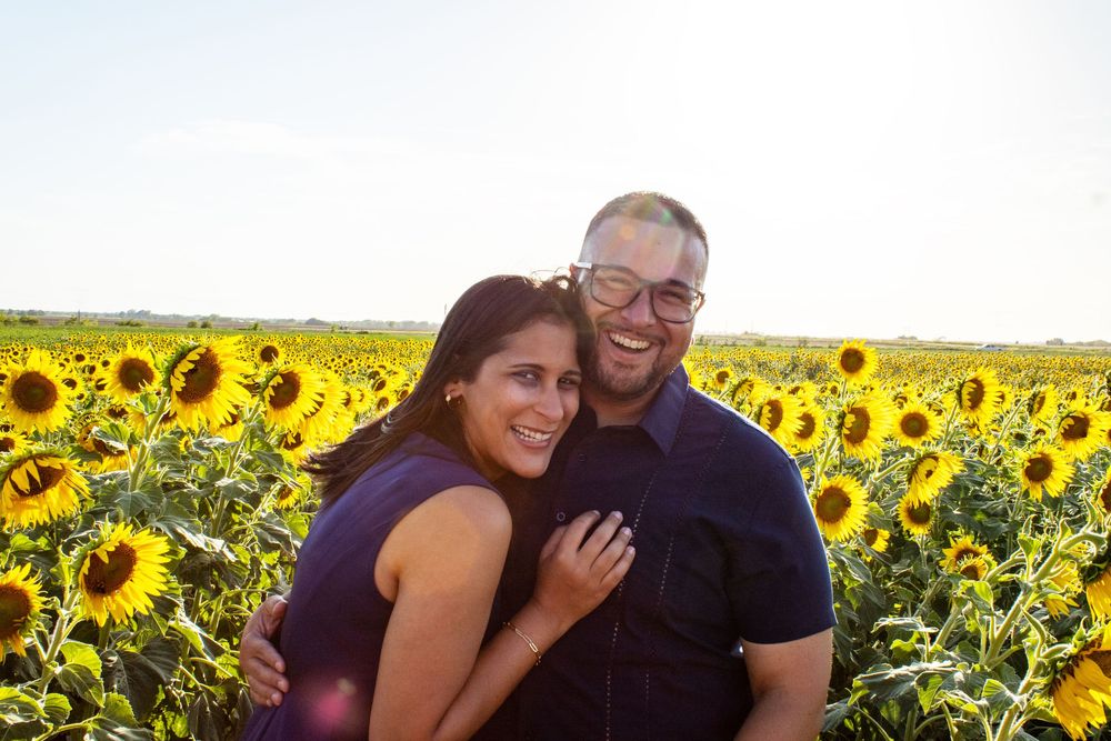 Couple smiling in a sunflower field on a sunny day, woman in purple dress embraced by man in blue shirt.